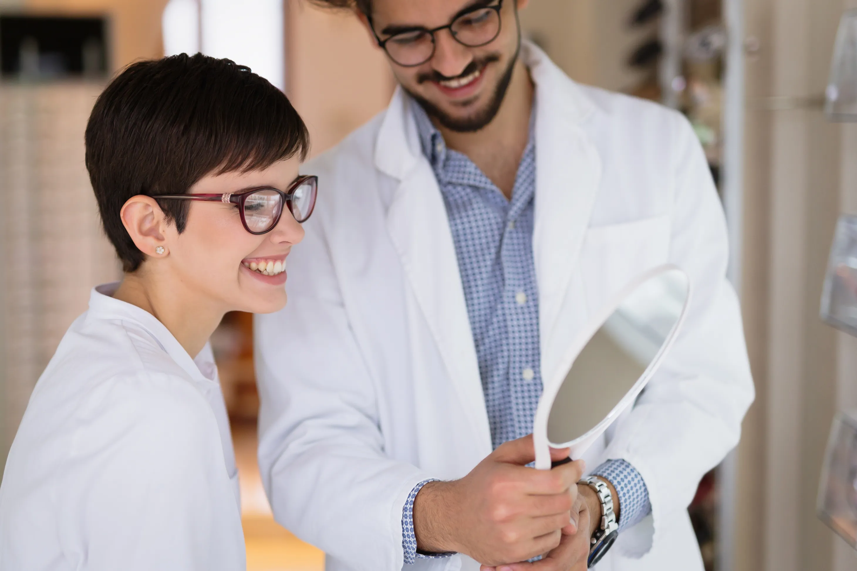 Smiling optician helping a woman try on eyeglasses while holding a hand mirror.