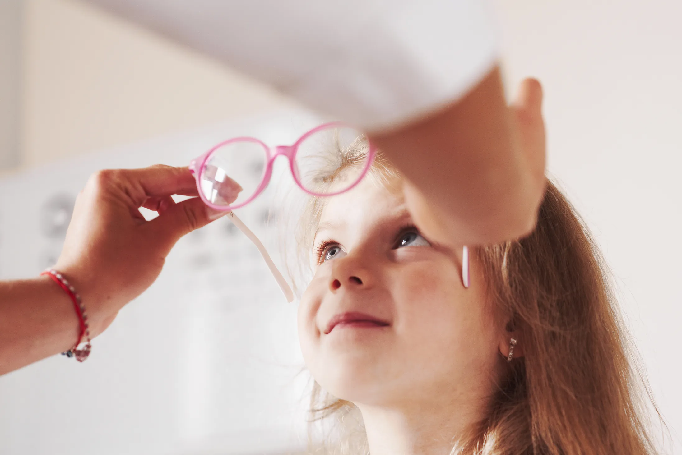 Close-up of a child looking up at pink eyeglasses being held by an adult.