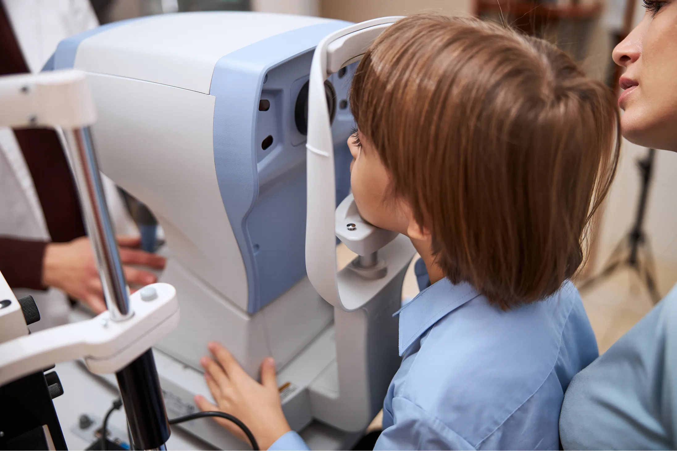 Child undergoing an eye exam using a digital autorefractor machine with an adult assisting.