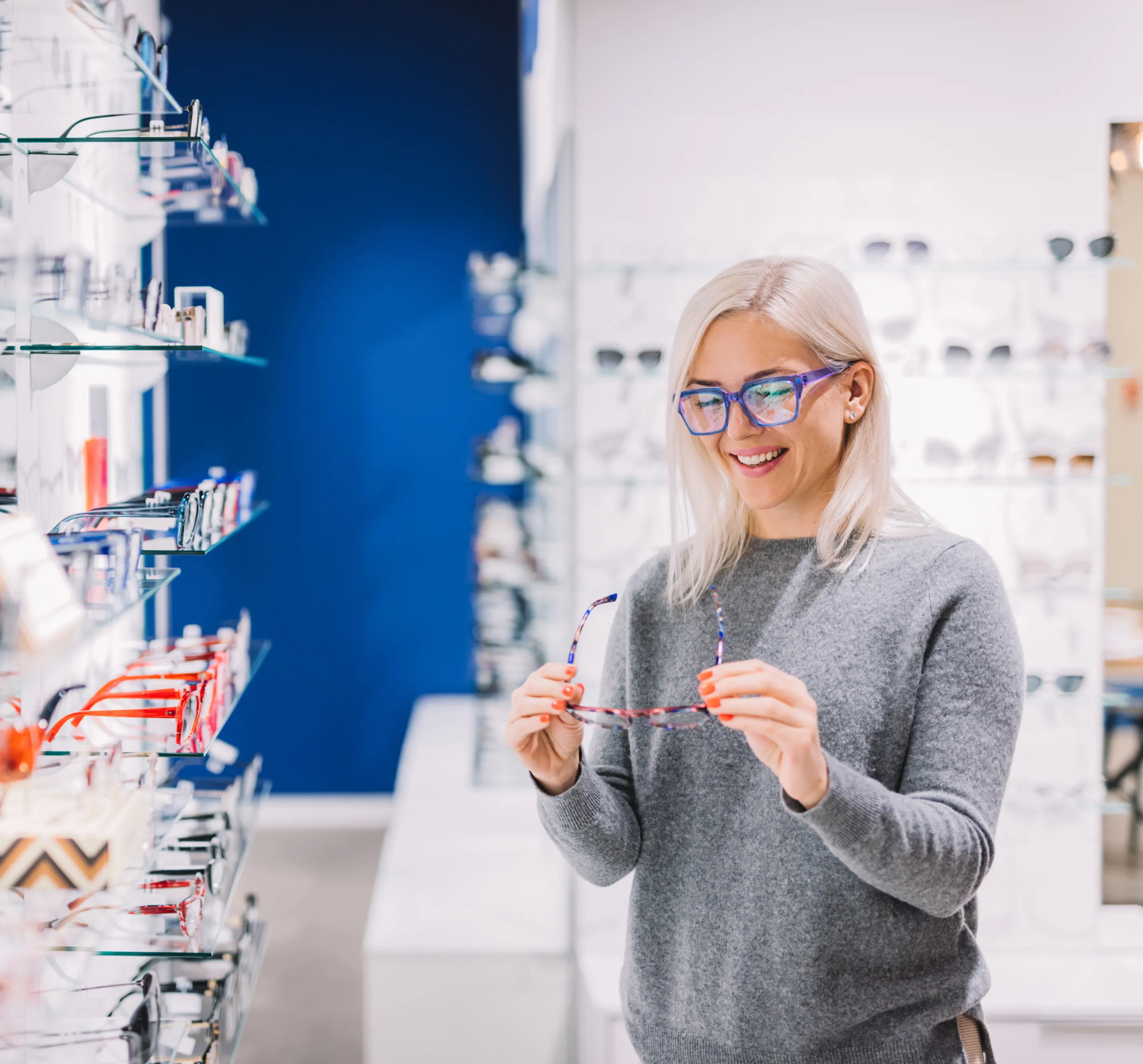Smiling woman with blonde hair wearing blue glasses while holding another pair of eyeglasses in an optical store.