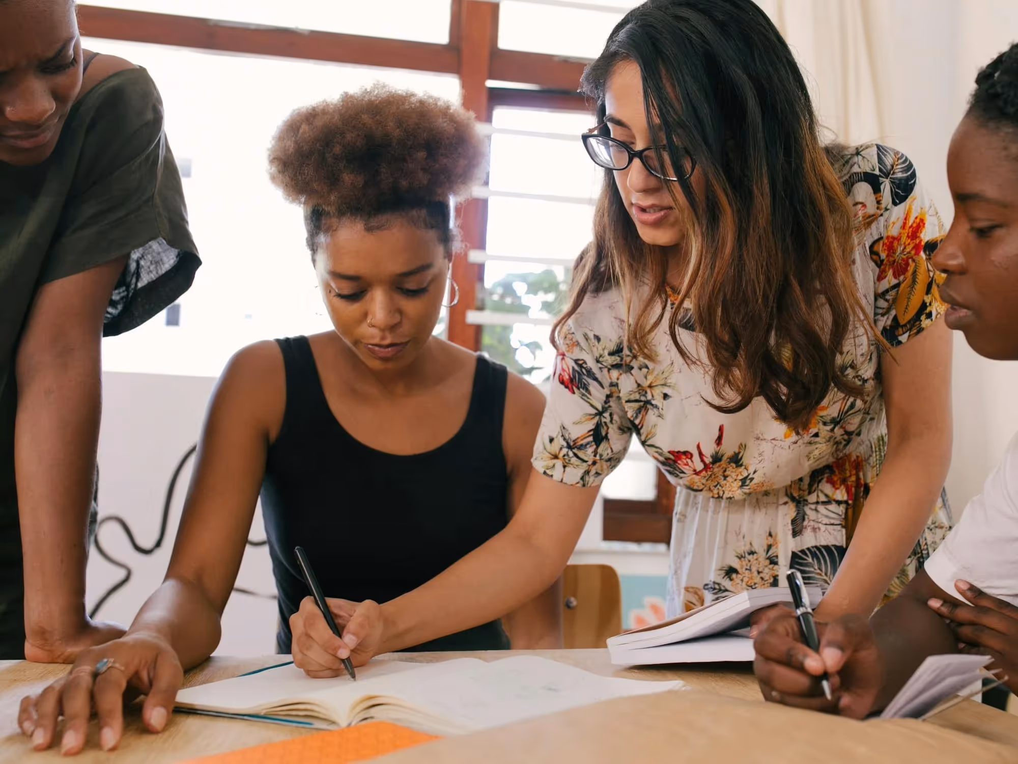 A group of four diverse young women studying together and writing in notebooks at a wooden table.