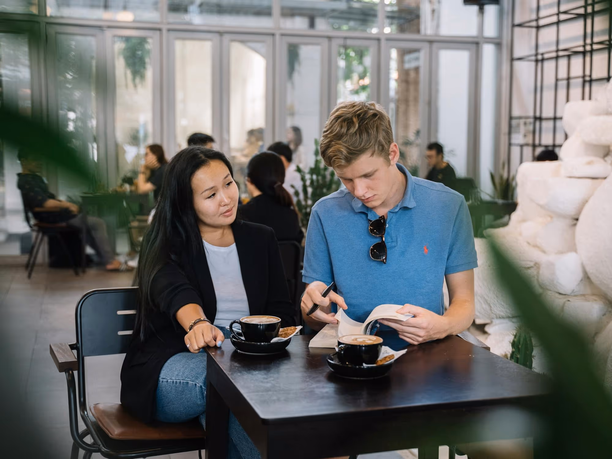 A young man reading a book and a woman looking at him, both seated at a café table with coffee cups.