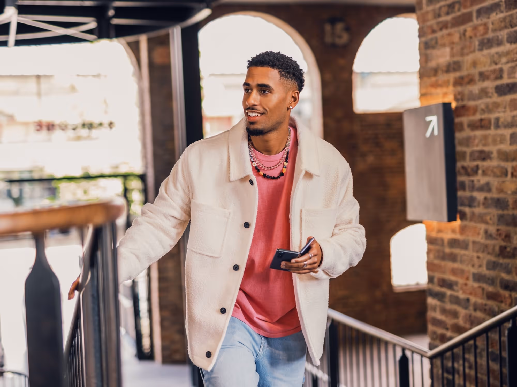Young man wearing a white jacket and pink shirt, holding a smartphone while walking up stairs indoors.