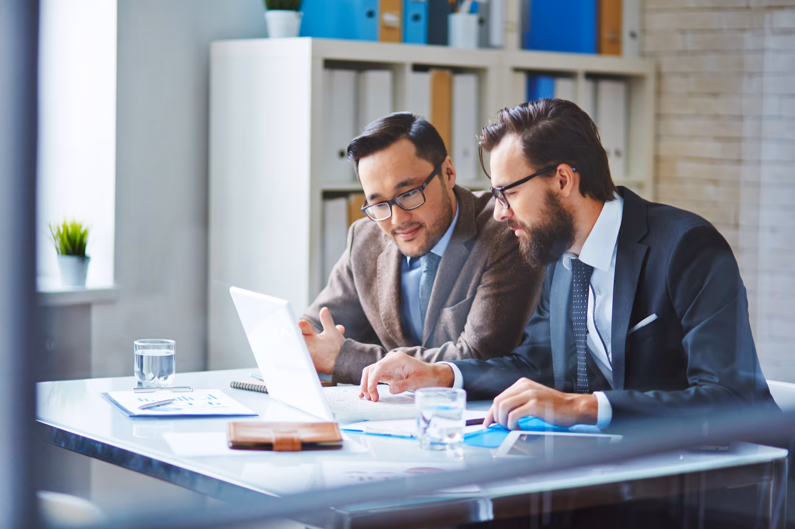Two businessmen in glasses sitting at a desk, looking at a laptop together in a modern office.