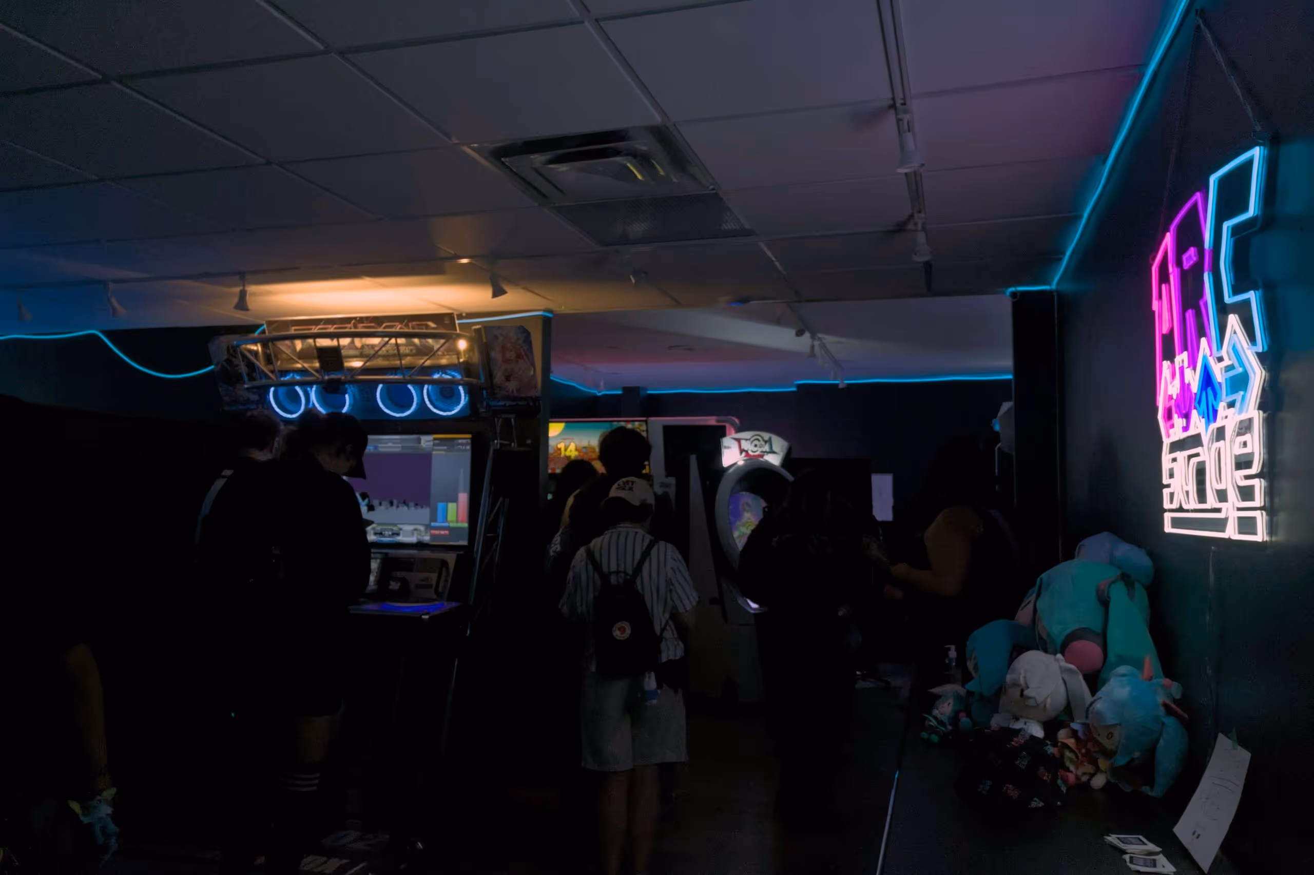 Dimly lit arcade with people playing games and a neon sign on the wall reading 'arcade' above shelves holding plush toys.