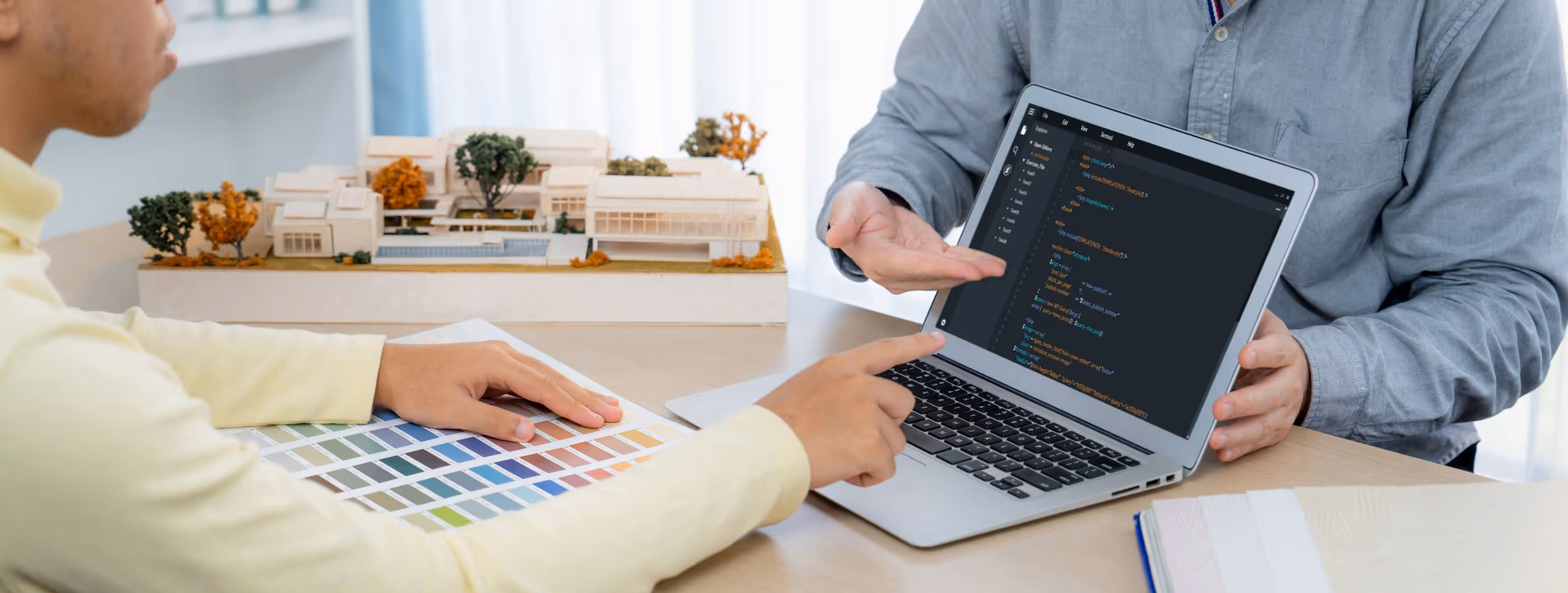 Two people discussing code on a laptop with a color palette and architectural model on the table.