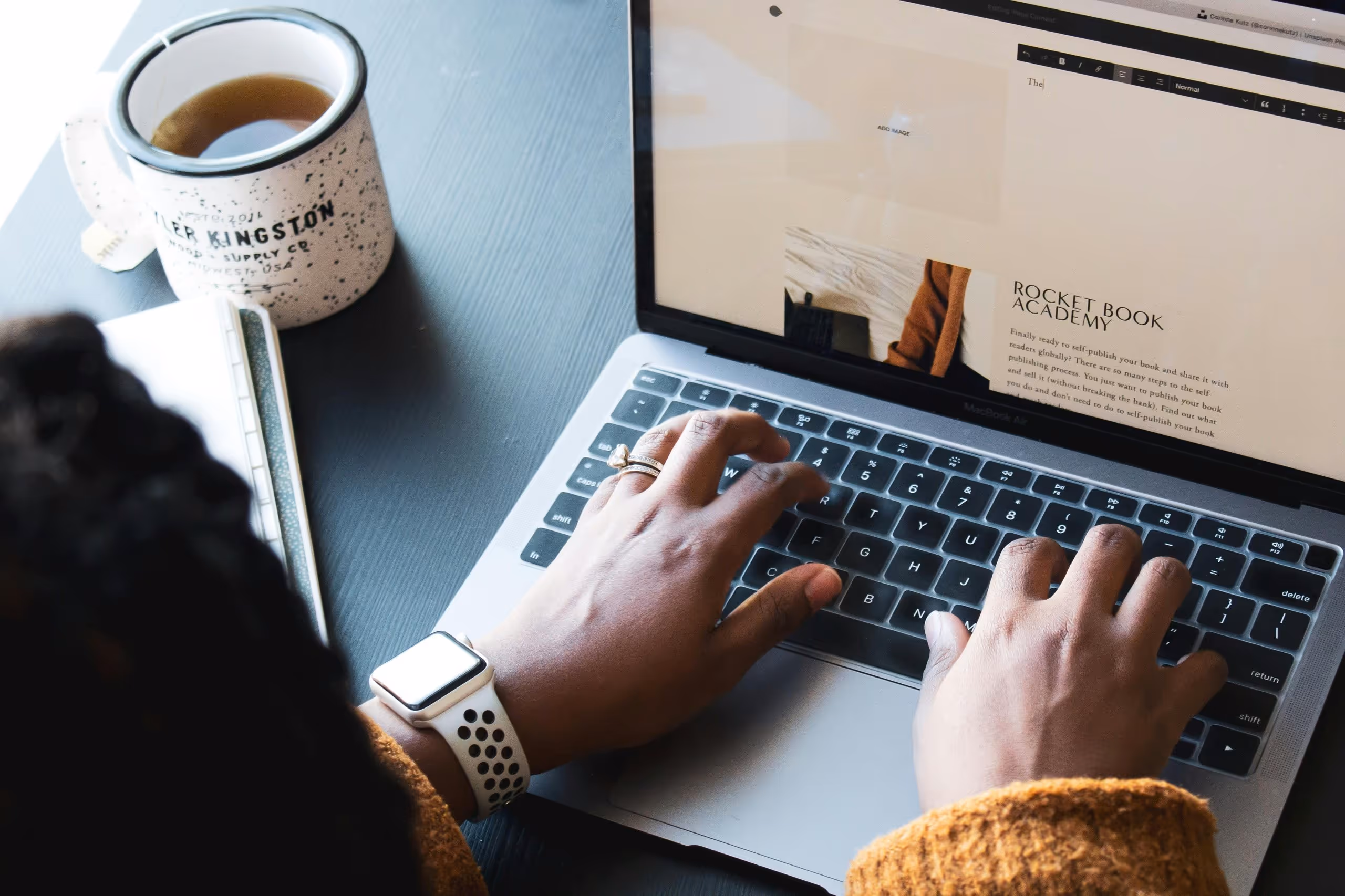 Person wearing a smartwatch and a ring typing on a MacBook Air keyboard with a cup of tea and a notebook on a dark table.