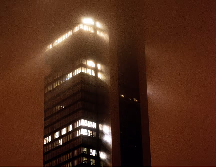 Tall modern building illuminated by interior lights at night with fog surrounding the upper floors.