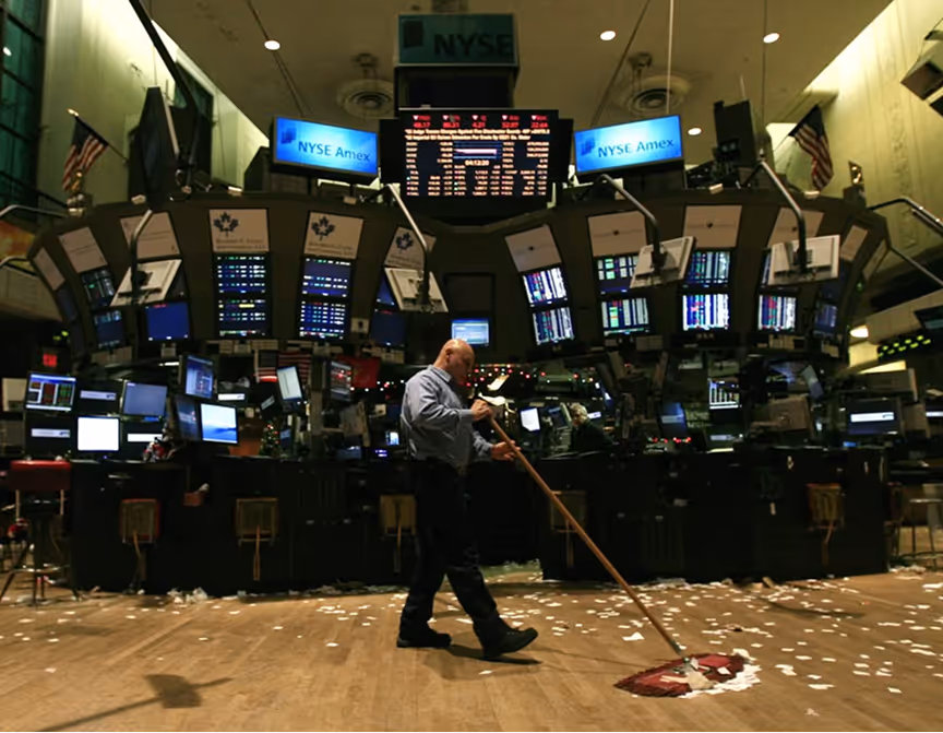 Person sweeping debris from the floor of the New York Stock Exchange trading floor with multiple screens and stock tickers in the background.