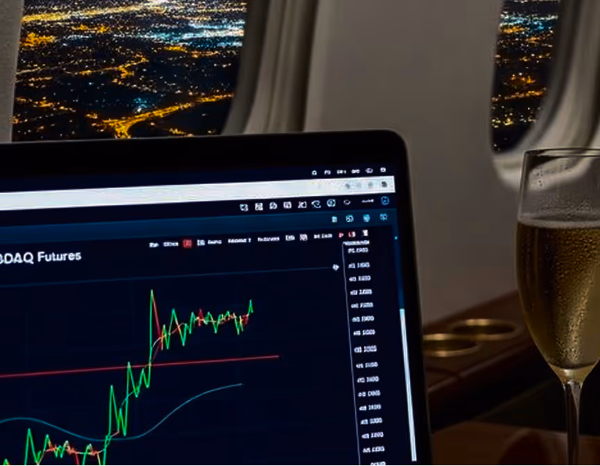 Laptop screen showing NASDAQ Futures stock chart inside an airplane, city lights visible through window, and a glass of champagne on a table.
