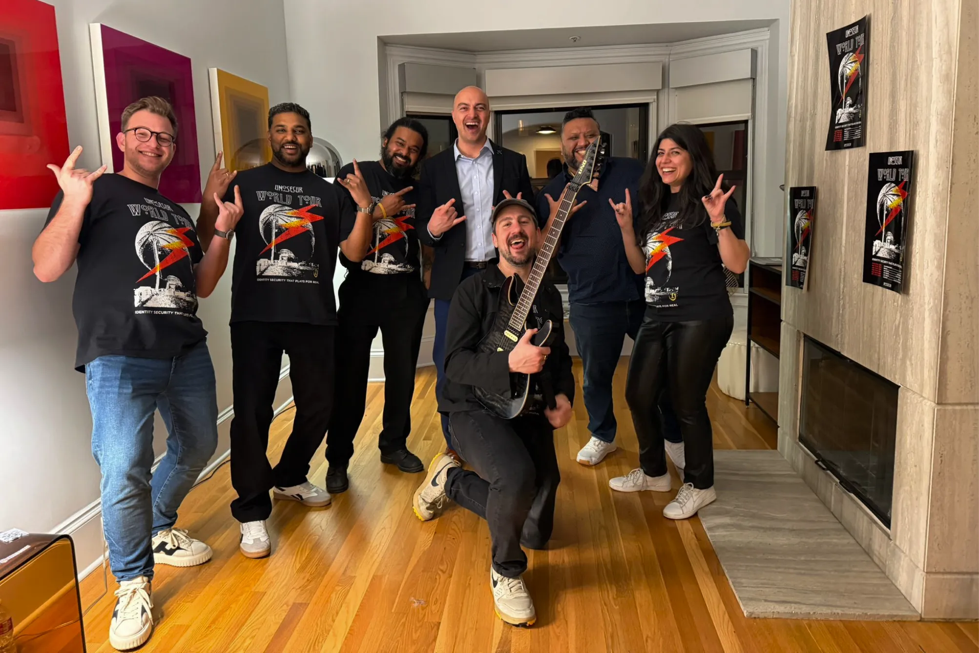 Group of seven people indoors smiling, five wearing matching black 'World Tour' shirts, one with a guitar.