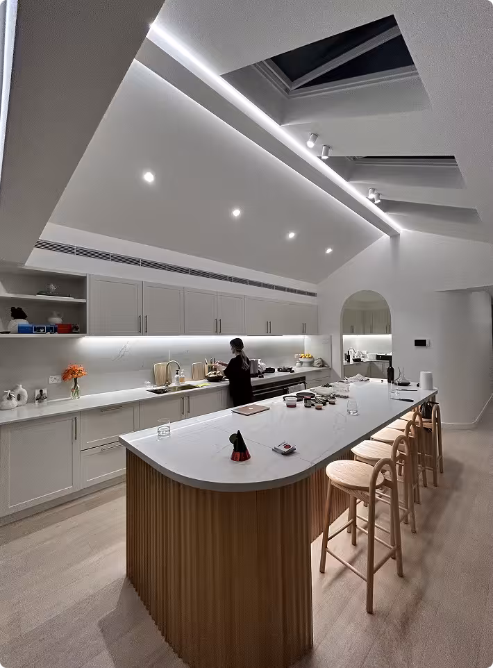 Modern kitchen with a sleek white island, wooden stools, under-cabinet lighting, and skylights; a person is preparing food at the counter.