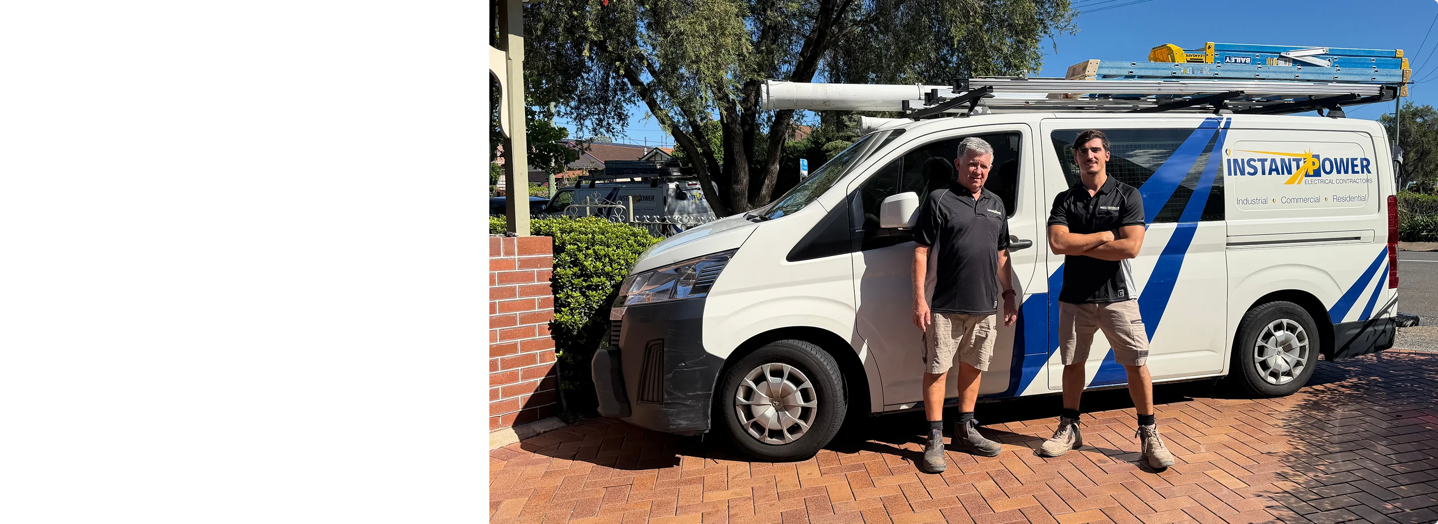 Two men in black shirts and tan shorts standing next to a white Instant Power electrical contractors van with ladders on the roof, parked on a brick driveway.