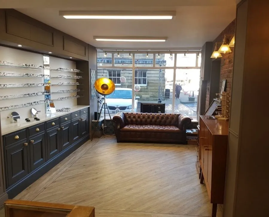 Interior of an eyewear store featuring a brown leather sofa, a large vintage floor lamp, and shelves displaying eyeglass frames.