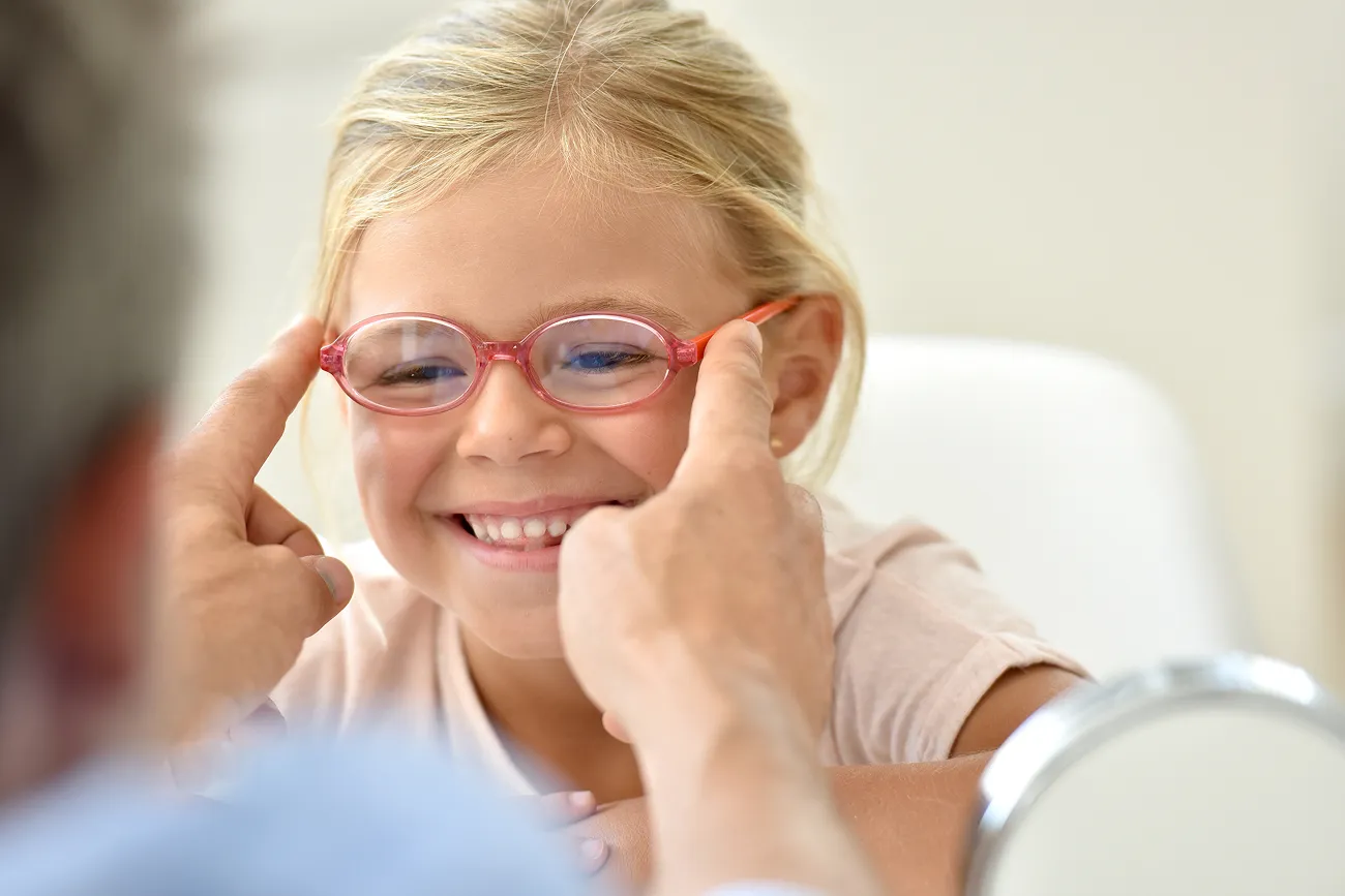 A smiling young girl with blonde hair tries on pink glasses being adjusted by an adult's hands.