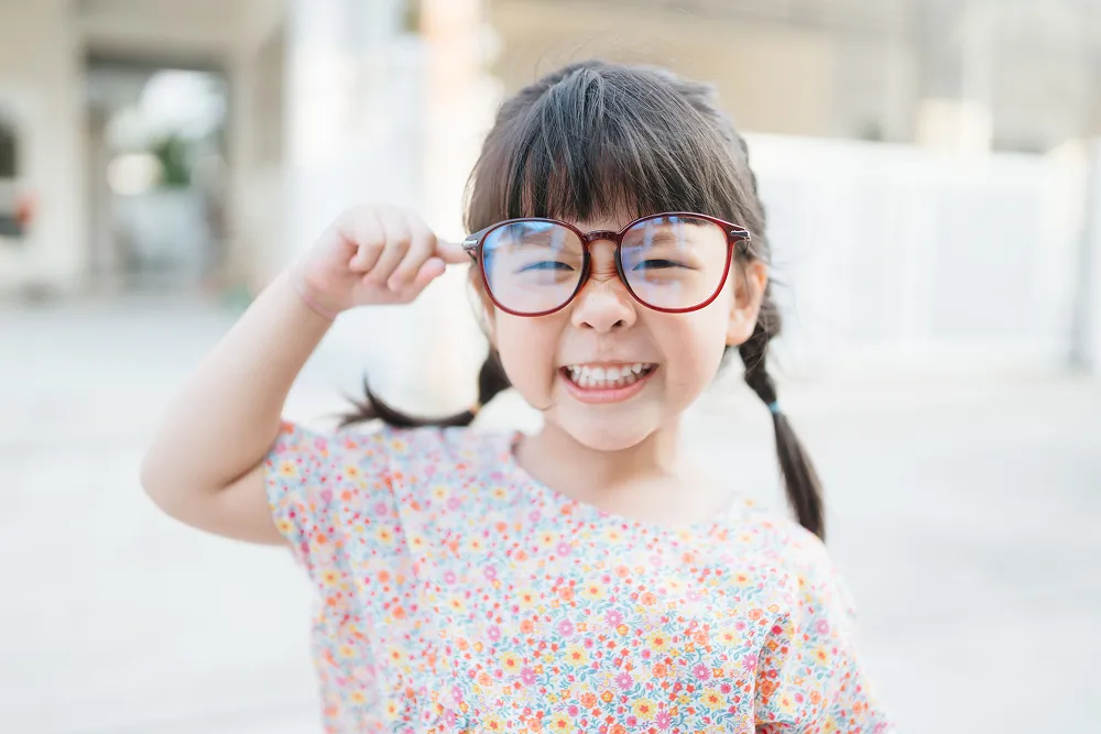 Smiling young girl with braided hair wearing oversized glasses and a floral shirt.