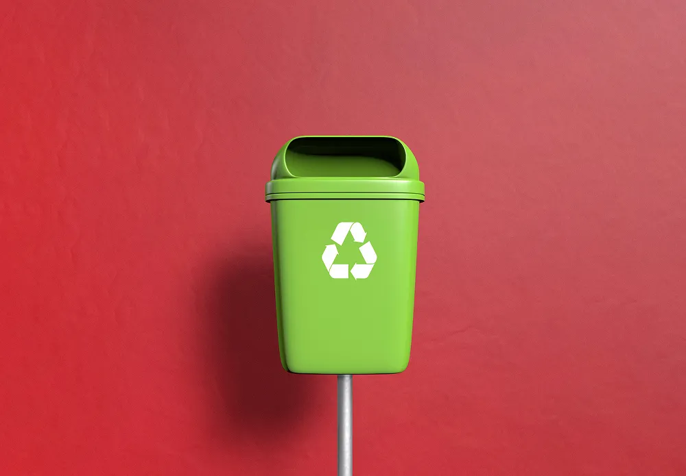 Green recycling bin with white recycling symbol mounted on a metal pole against a red wall.