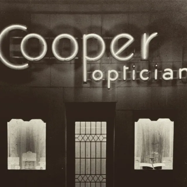 Black and white photo of a storefront sign reading 'Cooper opticians' with lighted letters above a door and two windows with curtains.