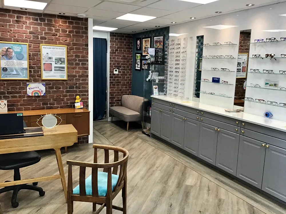 Interior of an optometry office with eyeglasses on display shelves, a wooden chair with a blue cushion, and brick accent walls.
