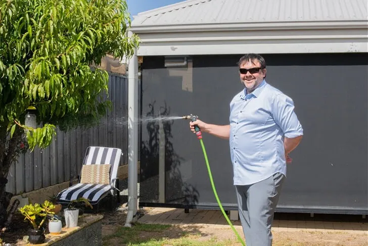 Man wearing sunglasses and a light blue shirt watering plants in a backyard with a green hose.