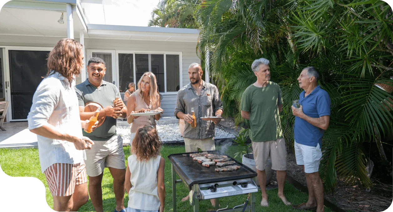 Group of adults and a child gathered outdoors around a grill with meat, enjoying a casual barbecue in a backyard with greenery.