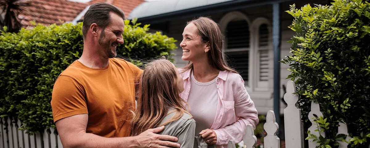 Smiling family of three embracing each other outdoors near a white picket fence and greenery.