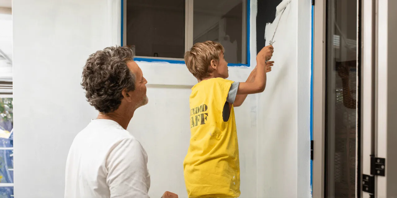 Man and young boy painting a wall white, with the boy using a paint roller near a window.