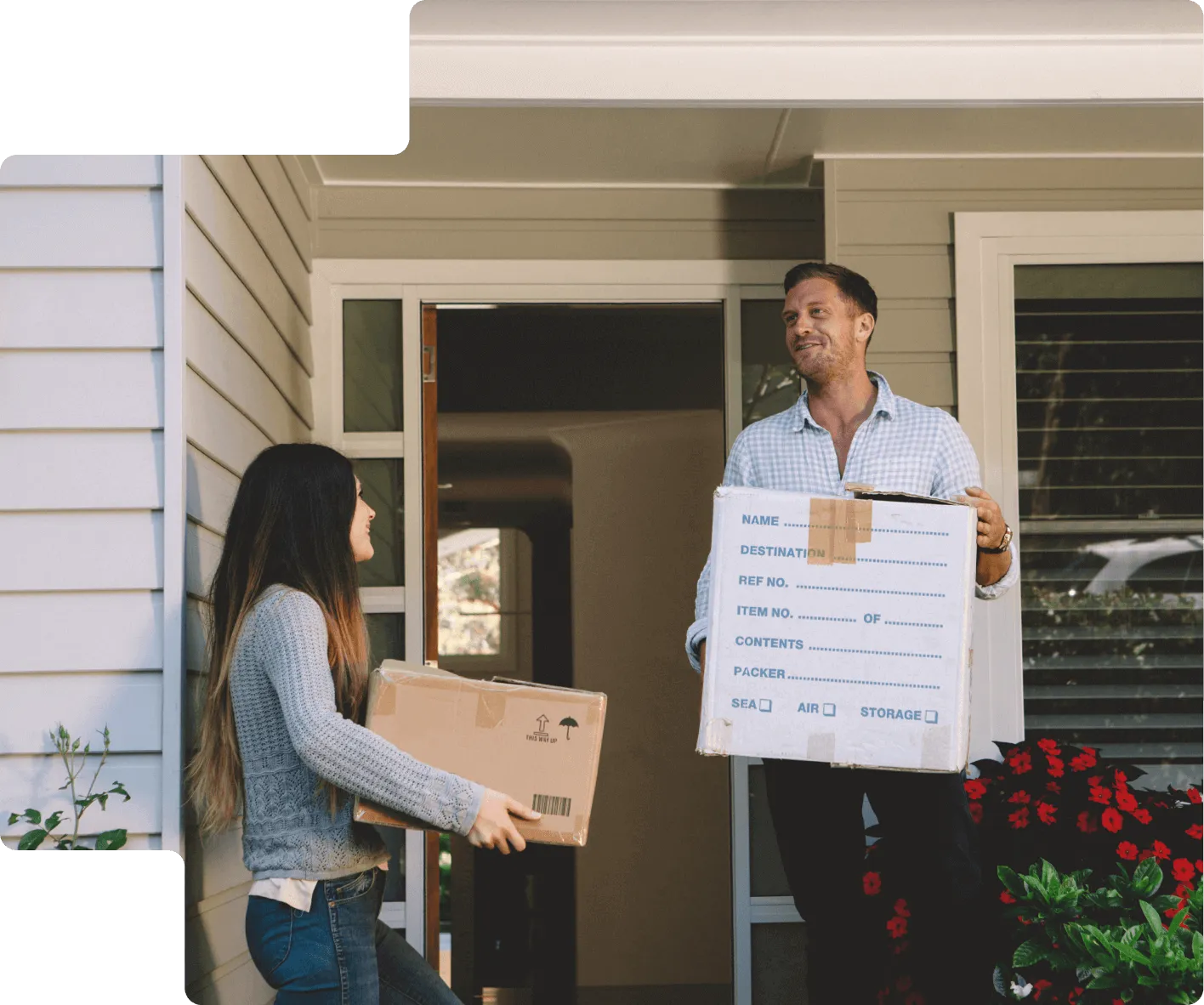 A man and a woman smiling and carrying cardboard boxes outside a house with grey siding and red flowers near the entrance.