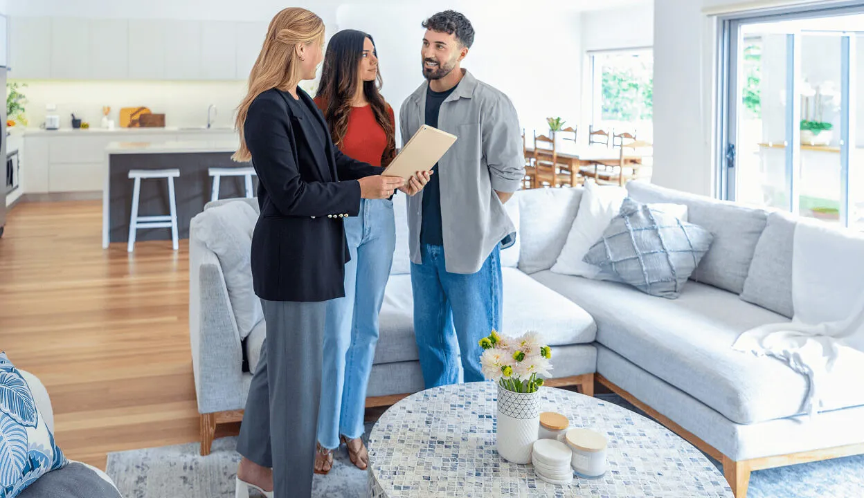 Real estate agent showing a tablet to a couple inside a bright, modern living room with light furniture and a round coffee table.