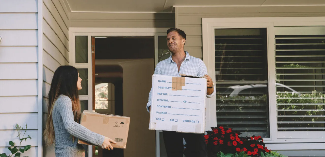 A man and woman carrying cardboard boxes while standing at the entrance of a house with gray siding and red flowers.