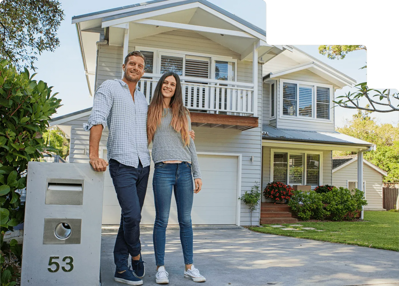 Smiling couple standing outside a modern two-story home with gray siding and a driveway, next to a mailbox with the number 53.