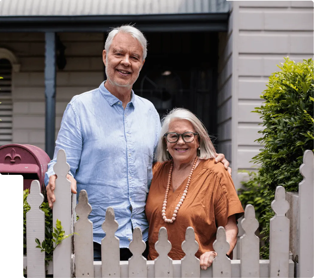 Smiling older couple standing behind a white picket fence outside their home.