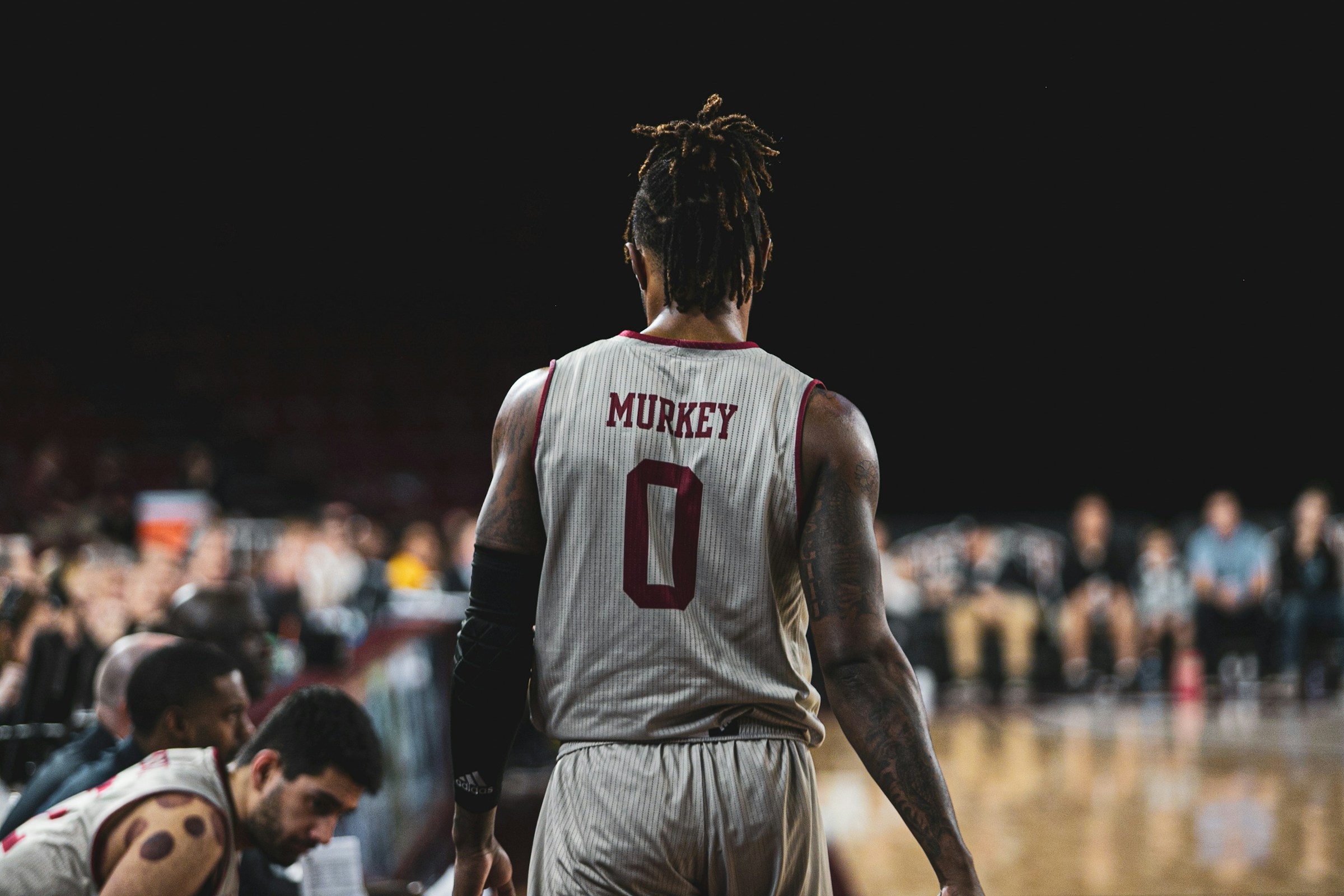 Basketball player standing on the court with their back to the camera during a game, surrounded by teammates and crowd under arena lights.