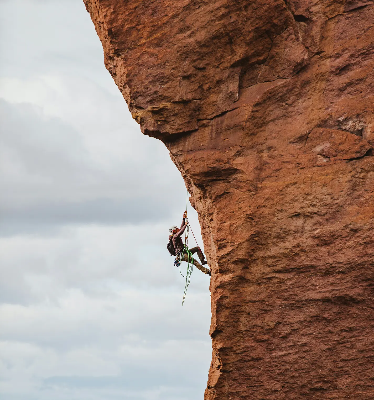 Jaxon Ruiz rock climbing a steep red sandstone cliff beneath an overhang, secured by rope against a cloudy sky.