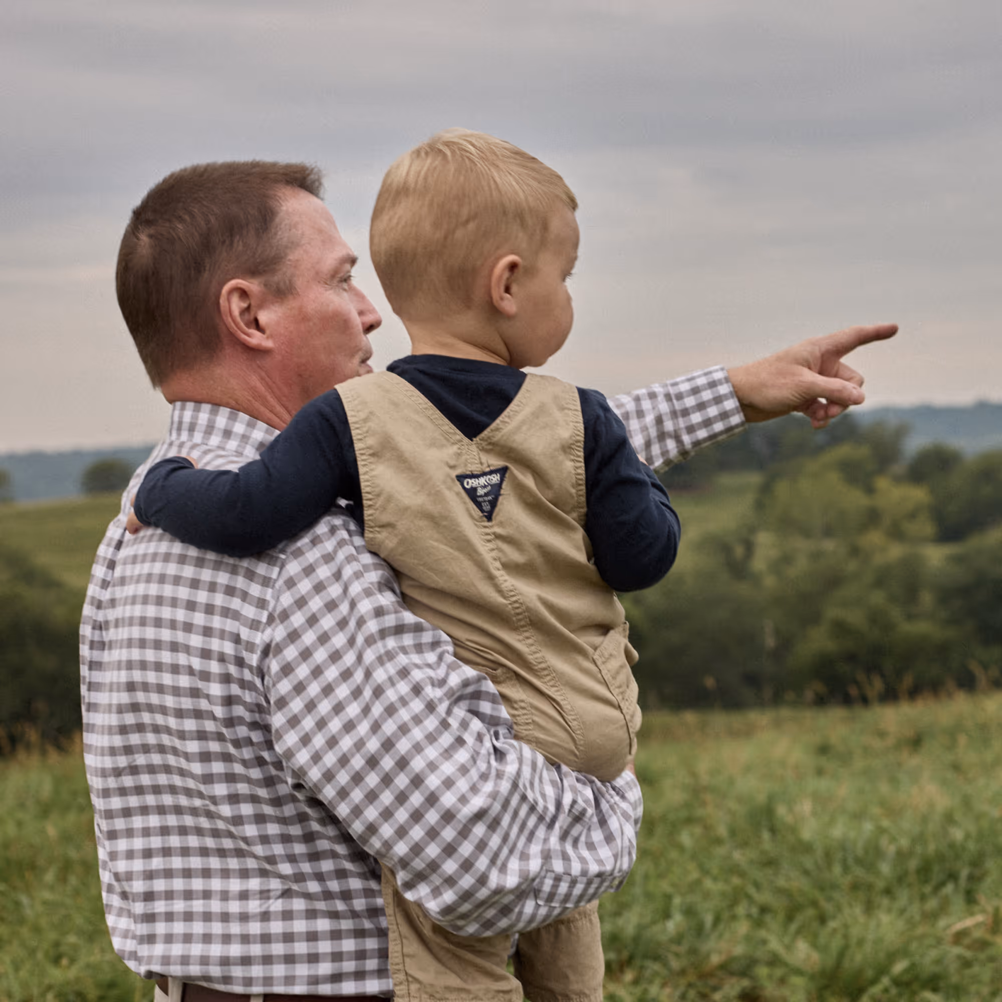 Man holding a young boy who is pointing towards something in a grassy field with trees in the background.