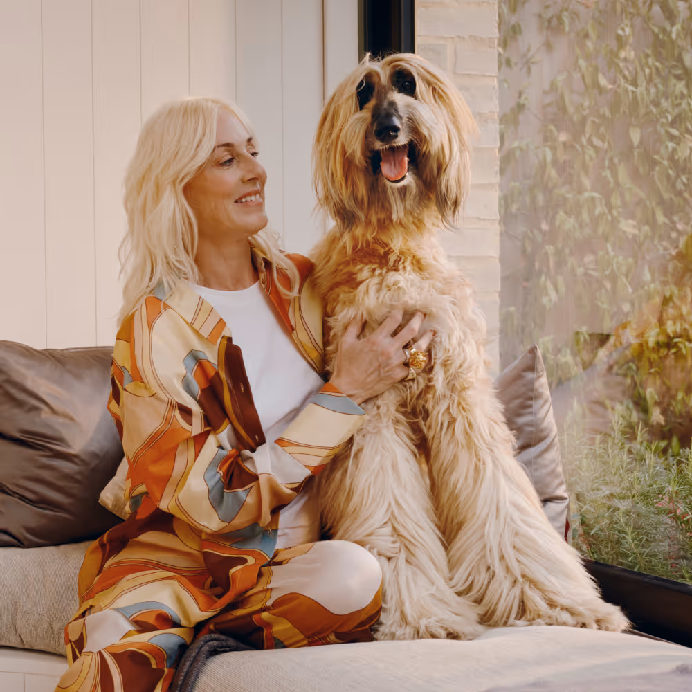 Blonde woman sitting on a couch holding a large, fluffy dog by a window with plants outside.