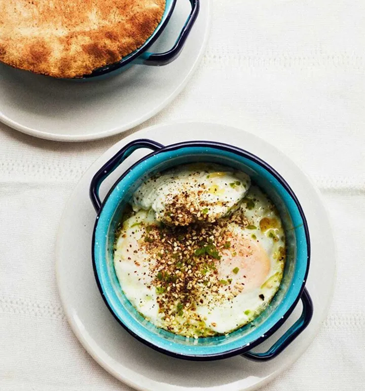 Two blue-handled pots on white plates; one contains baked eggs topped with seeds and herbs, the other has a golden-brown crust.