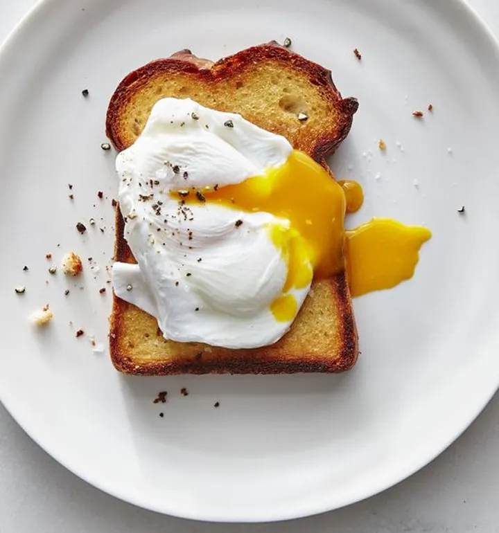Poached egg with runny yolk on a slice of toasted bread, sprinkled with ground black pepper on a white plate.
