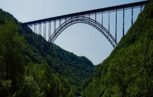 Steel arch bridge spanning a deep forested valley with clear sky above.