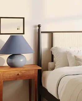 Cozy bedroom corner with a wooden nightstand holding a blue lamp beside a bed with white pillows and beige bedding.