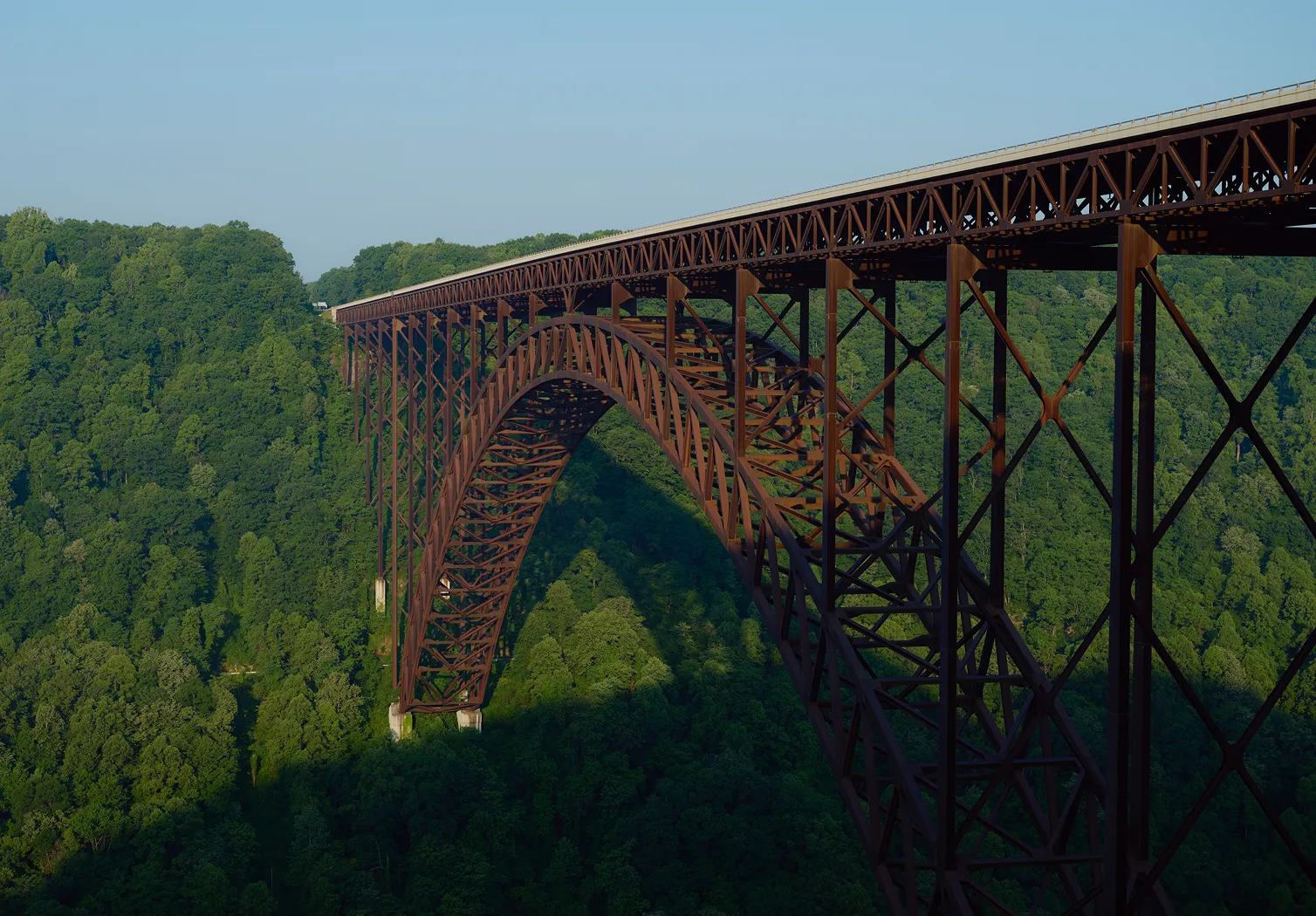 A large steel arch bridge spanning a deep green forested valley under a clear blue sky.