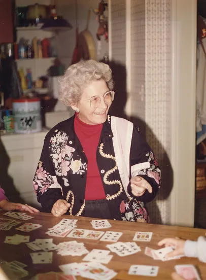 Elderly woman with curly gray hair and glasses smiling while seated at a table covered with playing cards.