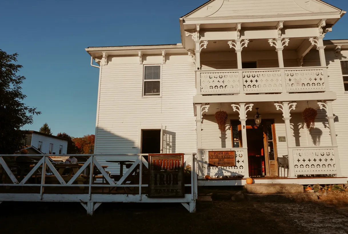 Two-story white wooden building with a porch decorated with hanging plants and pumpkins, under a clear blue sky.