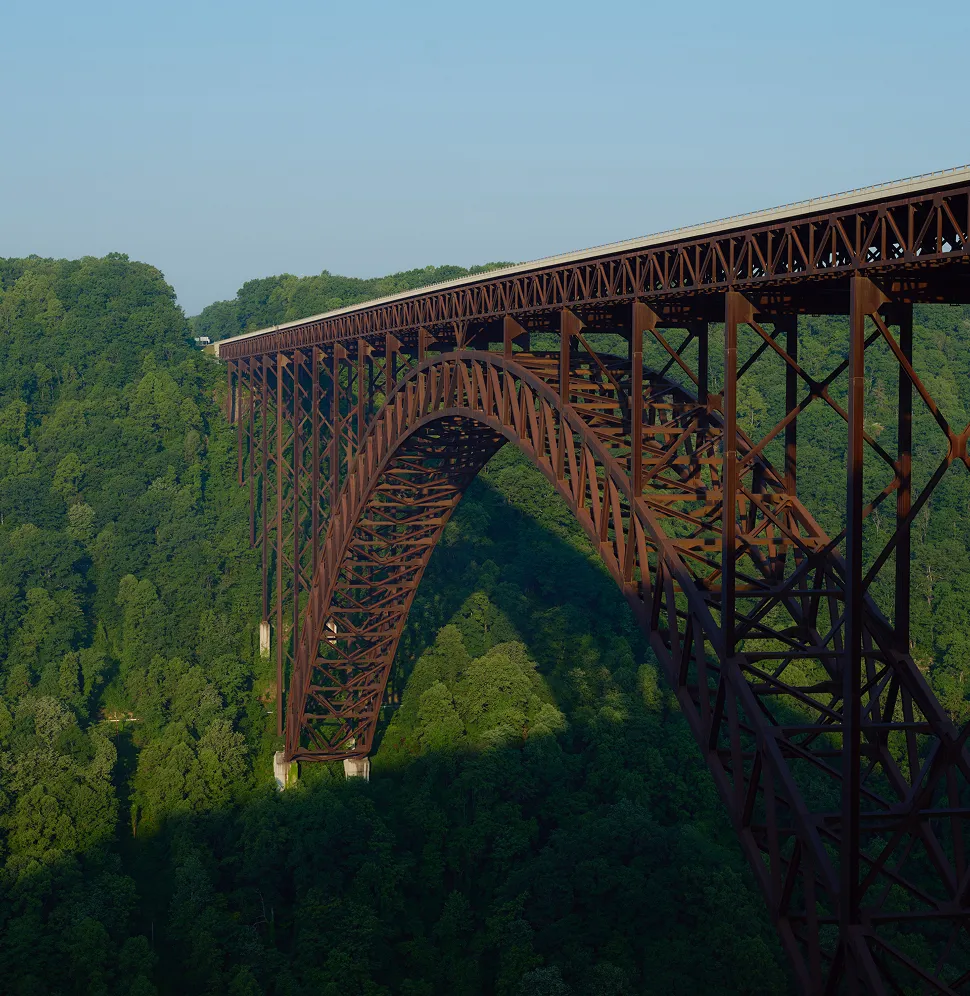 Rust-colored steel arch bridge spanning a deep green forest valley under a clear blue sky.