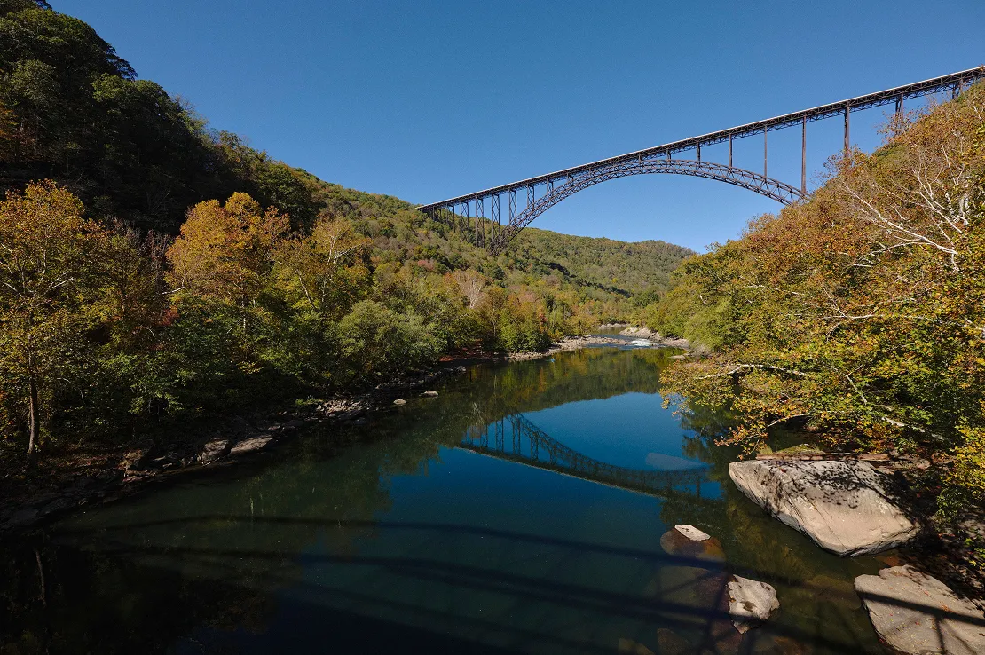 Iron arch bridge spanning over a calm river surrounded by autumn-colored trees under a clear blue sky.