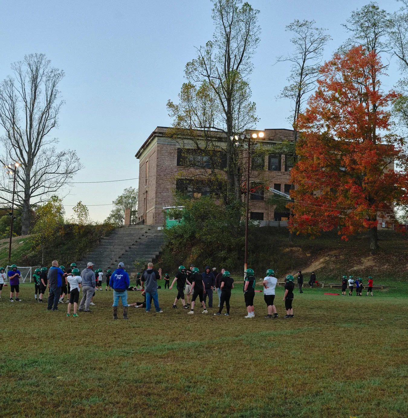 Group of youth football players wearing green helmets practicing on a grassy field near a building with autumn-colored trees.