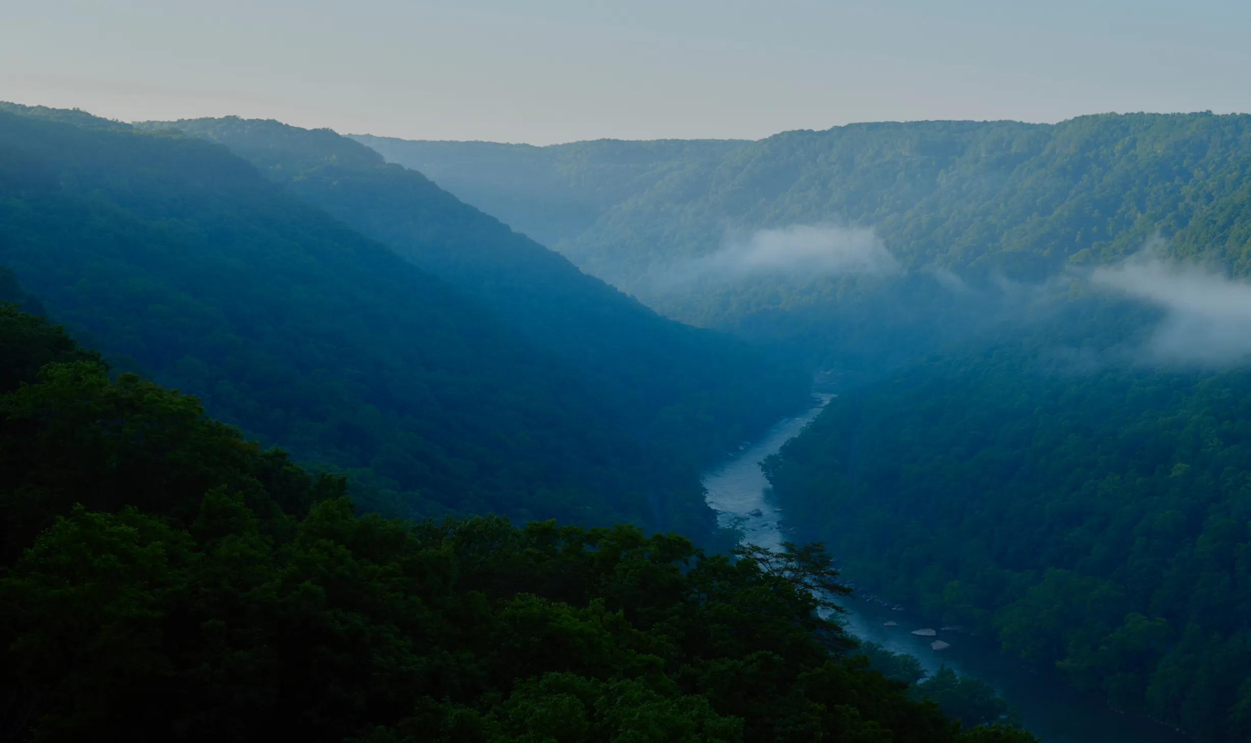 A river winding through lush green forested mountains under a clear sky with wisps of mist.