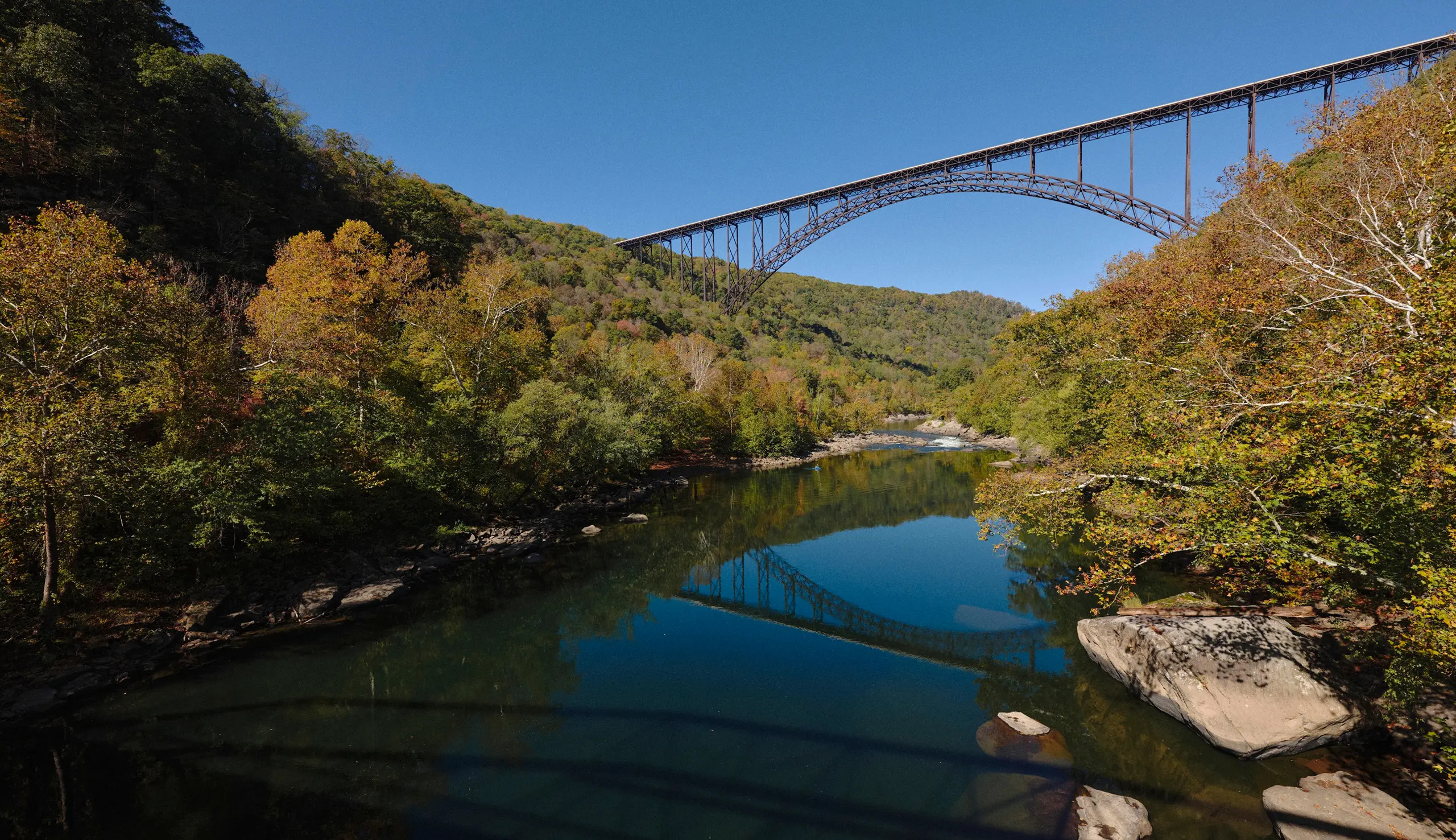 Steel arch bridge spanning over a calm river surrounded by autumn trees under a clear blue sky.