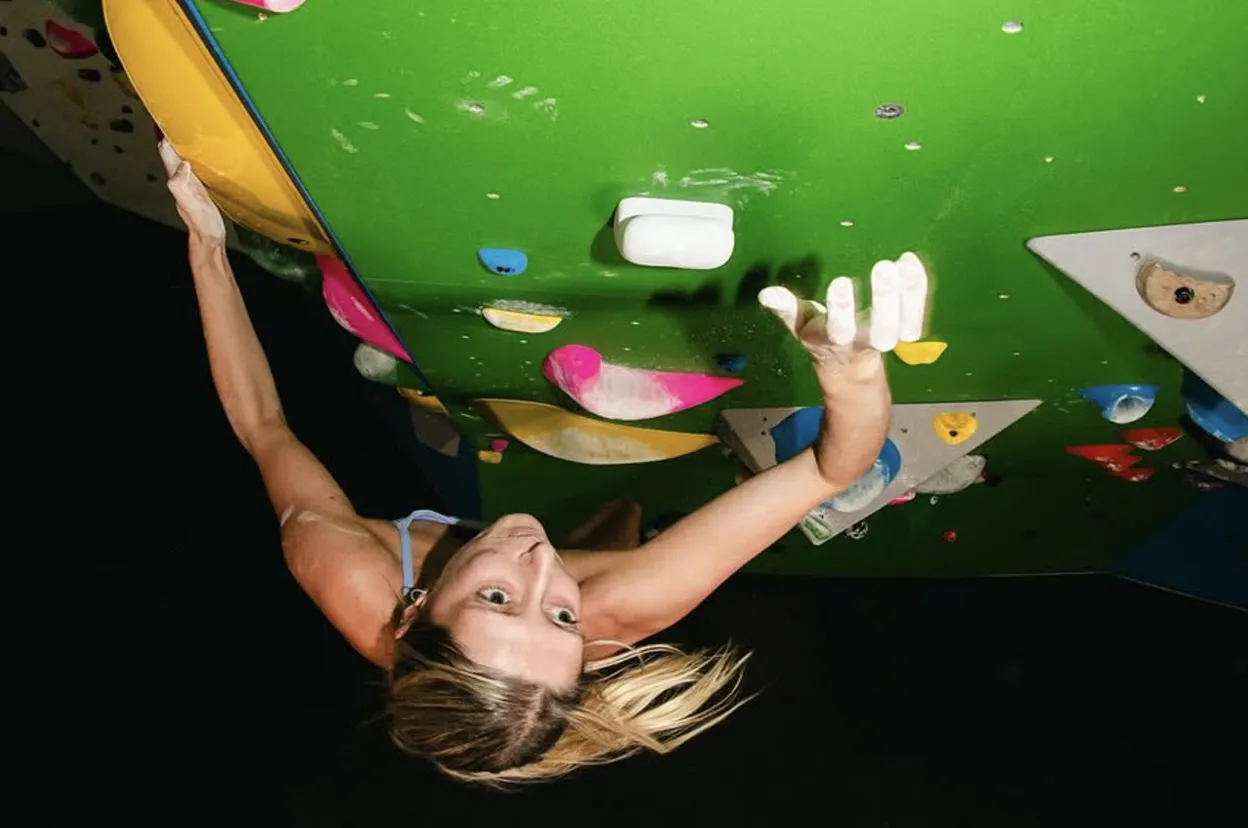 Woman climbing a green indoor rock climbing wall with colorful holds, reaching up with chalked hands.