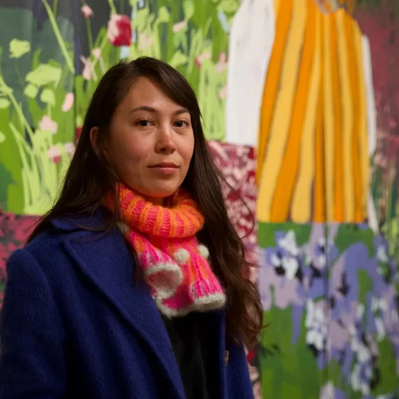 Woman wearing a blue coat and a colorful knitted scarf standing in front of a vibrant, floral mural.
